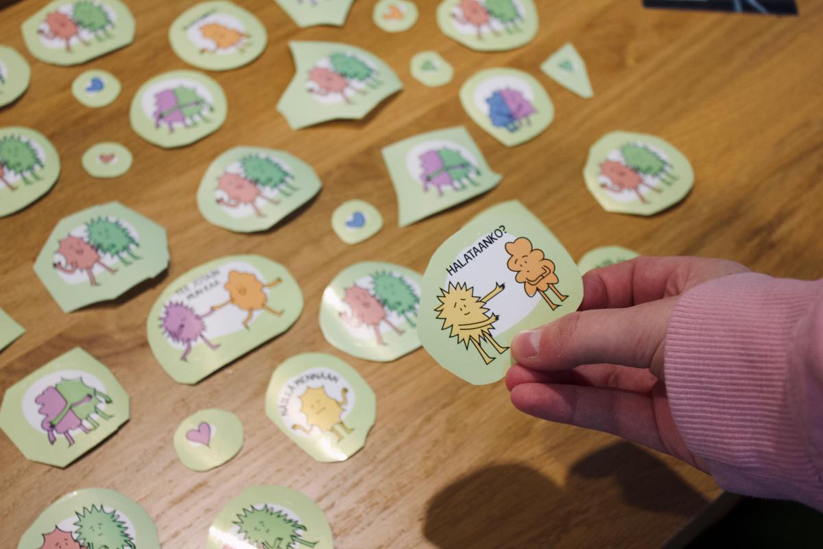 Pöydällä kuvallisia lappuja, jotka kuvaavat erilaisia kohtaamisia, etualalla henkilön kädessä lappu, jossa lukee: 