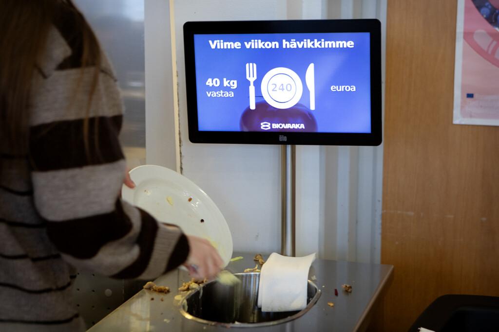 Ihminen laittamassa ruokaa roskikseen koulun ruokailussa. Taustalla näyttö, jossa lukee "Viime viikon hävikkimme 40kg vastaa 240 euroa - Biovaaka".