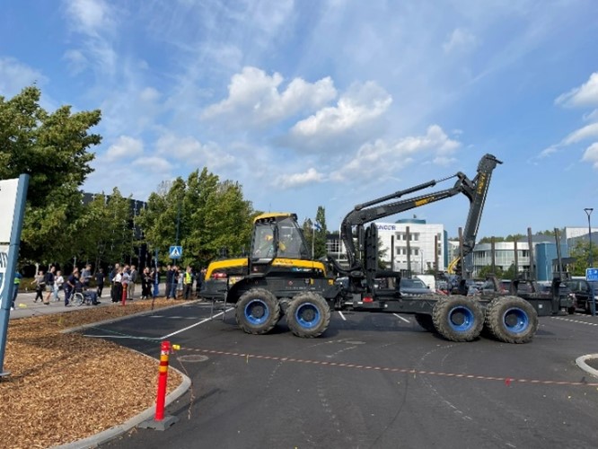 Iso keltamusta työkone parkkipaikalla, ihmisiä ympärillä, kaunis kesäpäivä.