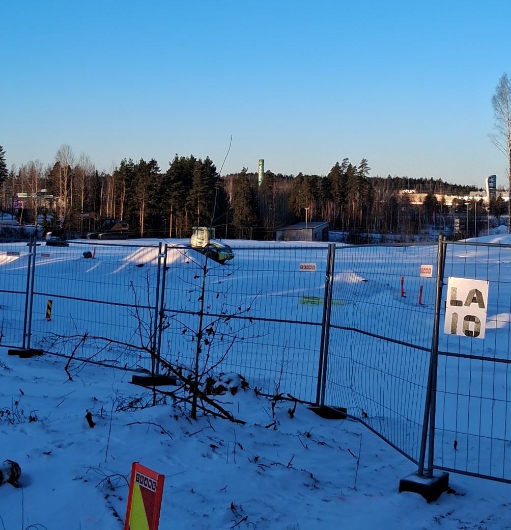 Työmaa-aidan takana tyhjä kenttä lumen alla aurinkoisena talvipäivänä.