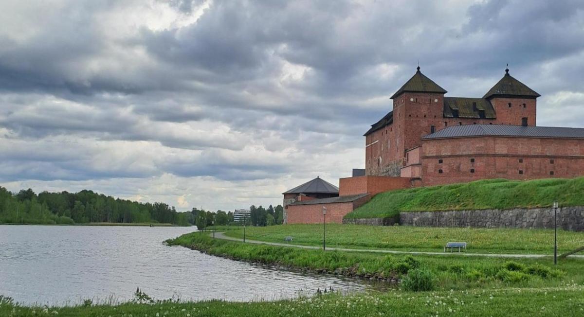 Kuvassa näkyy osa Hämeen linnasta sekä maisemaa järvestä ja pilvisestä taivaasta.