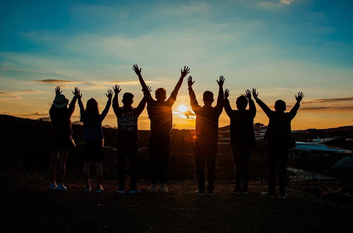 Aurinkoa vasten otettu kuva, jossa rivissä joukko ihmisiä käsi kädessä ja kädet kohti taivasta.